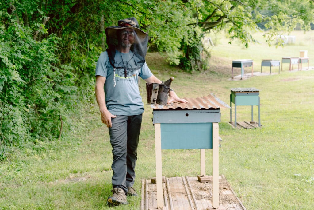 Beekeeper wearing a veil standing beside a small hive box in a grassy field