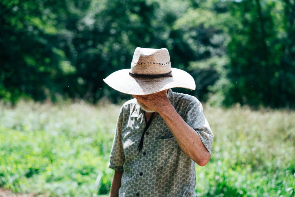 Farmer in straw hat standing in a grassy field with hand covering his face in frustration