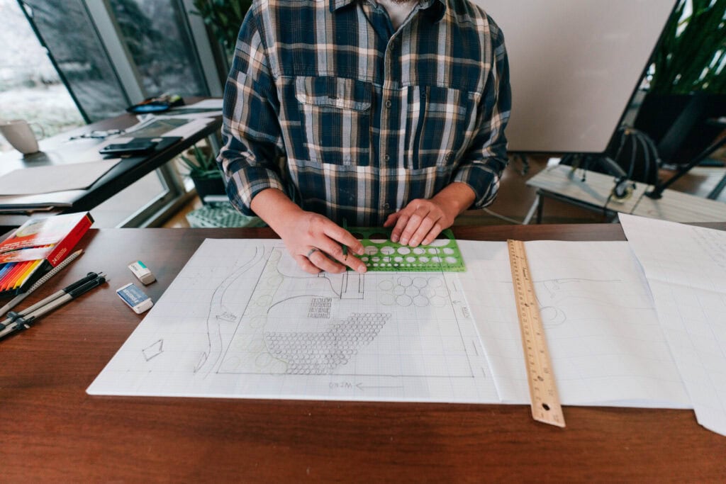 Person using a stencil and ruler to design a homestead layout on graph paper at a desk