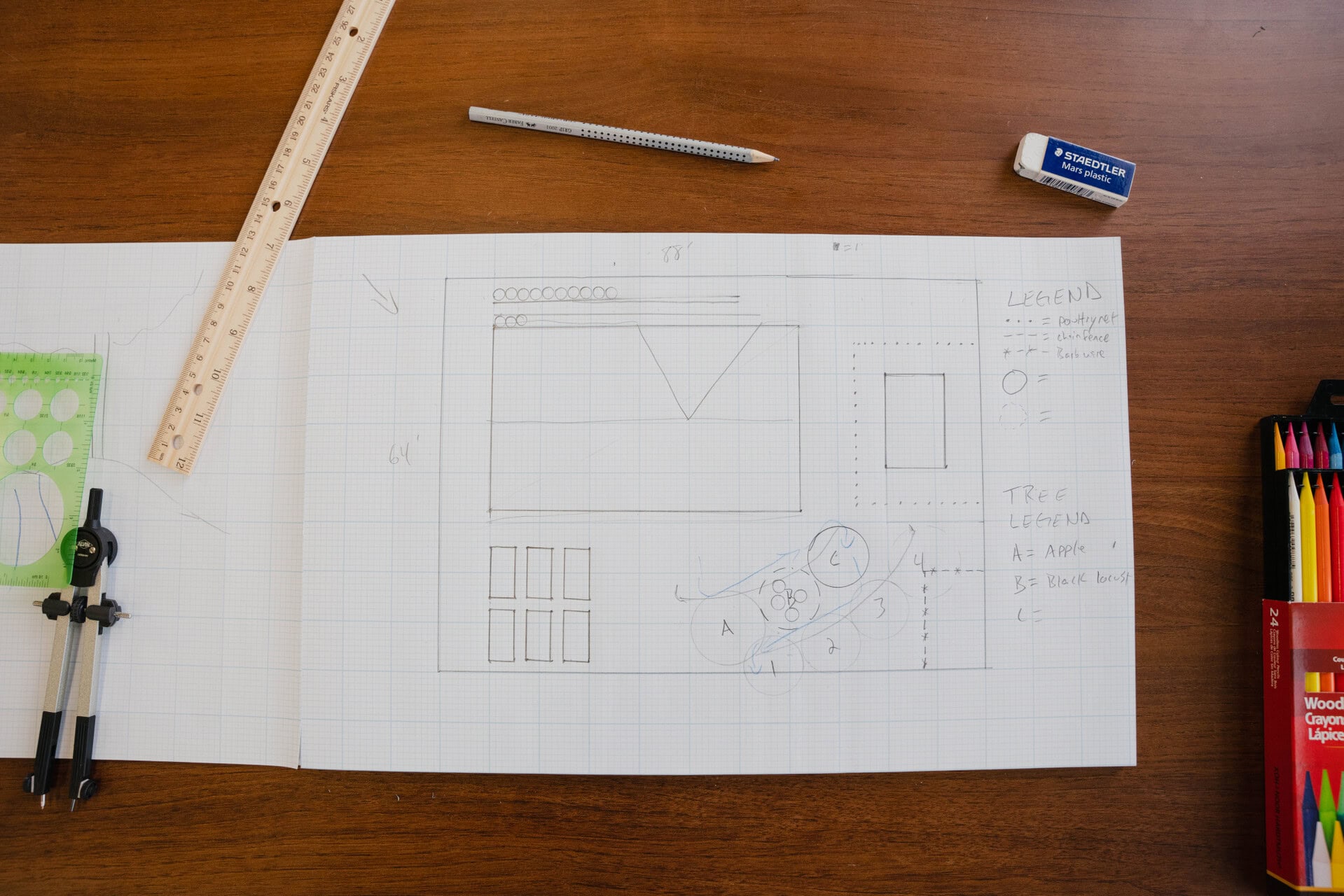 Homestead layout sketch on graph paper surrounded by ruler, pencils, and drawing tools on a wooden table