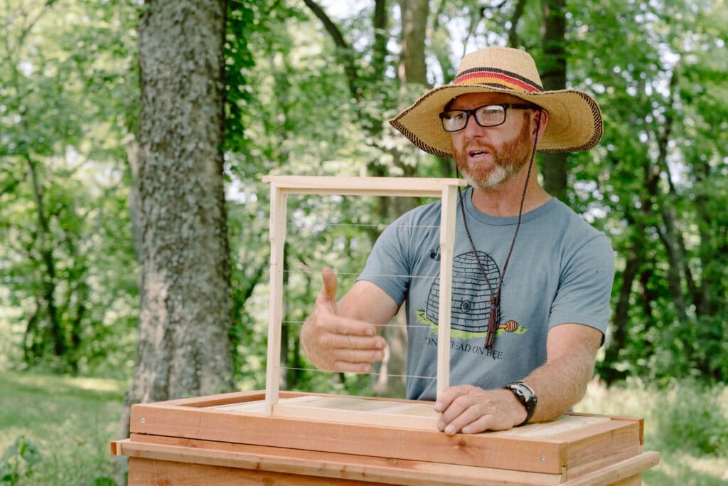 Beekeeper demonstrating a wooden hive frame with guide wires for natural comb building outdoors