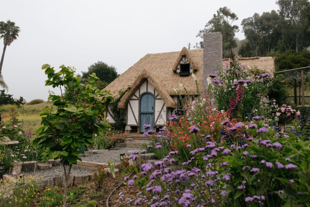 Diverse cottage garden with flowers, shrubs, and a thatched-roof home showing companion planting in a layered landscape
